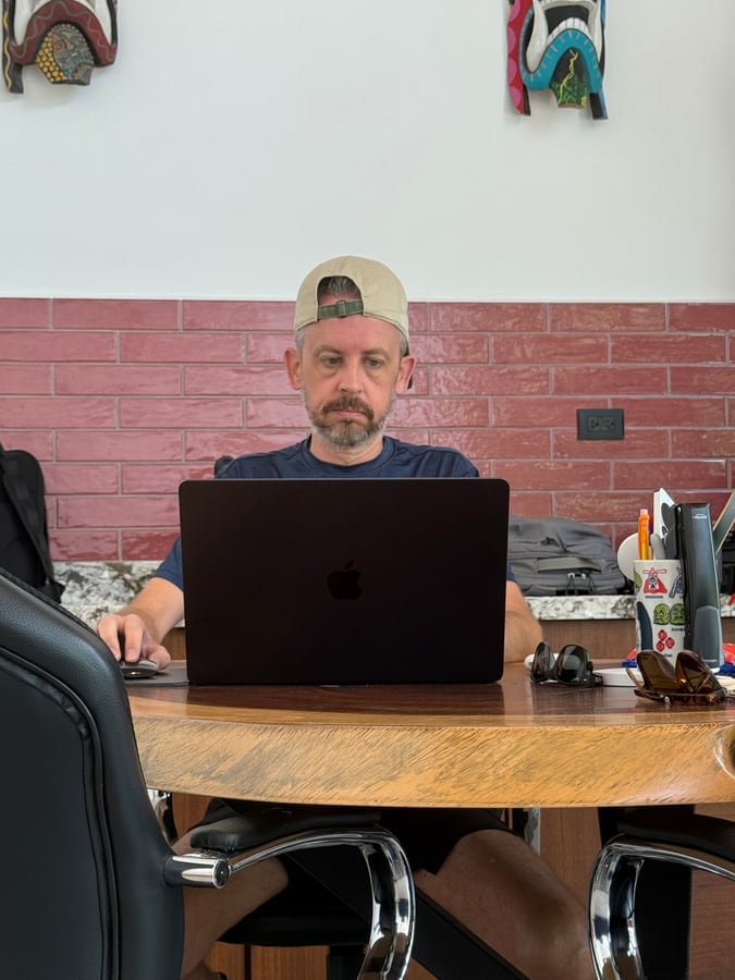 Working at a laptop at a round wooden table, masks on the wall behind